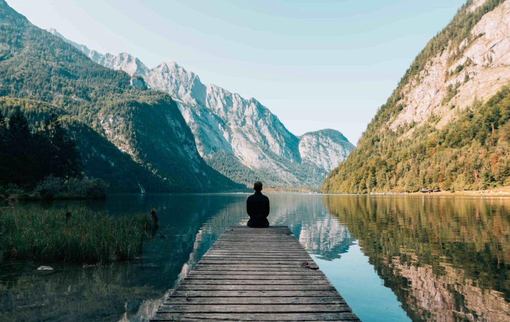 A person sits on the end of a deck jutting out into a serene lake looking onto a forested mountain range.